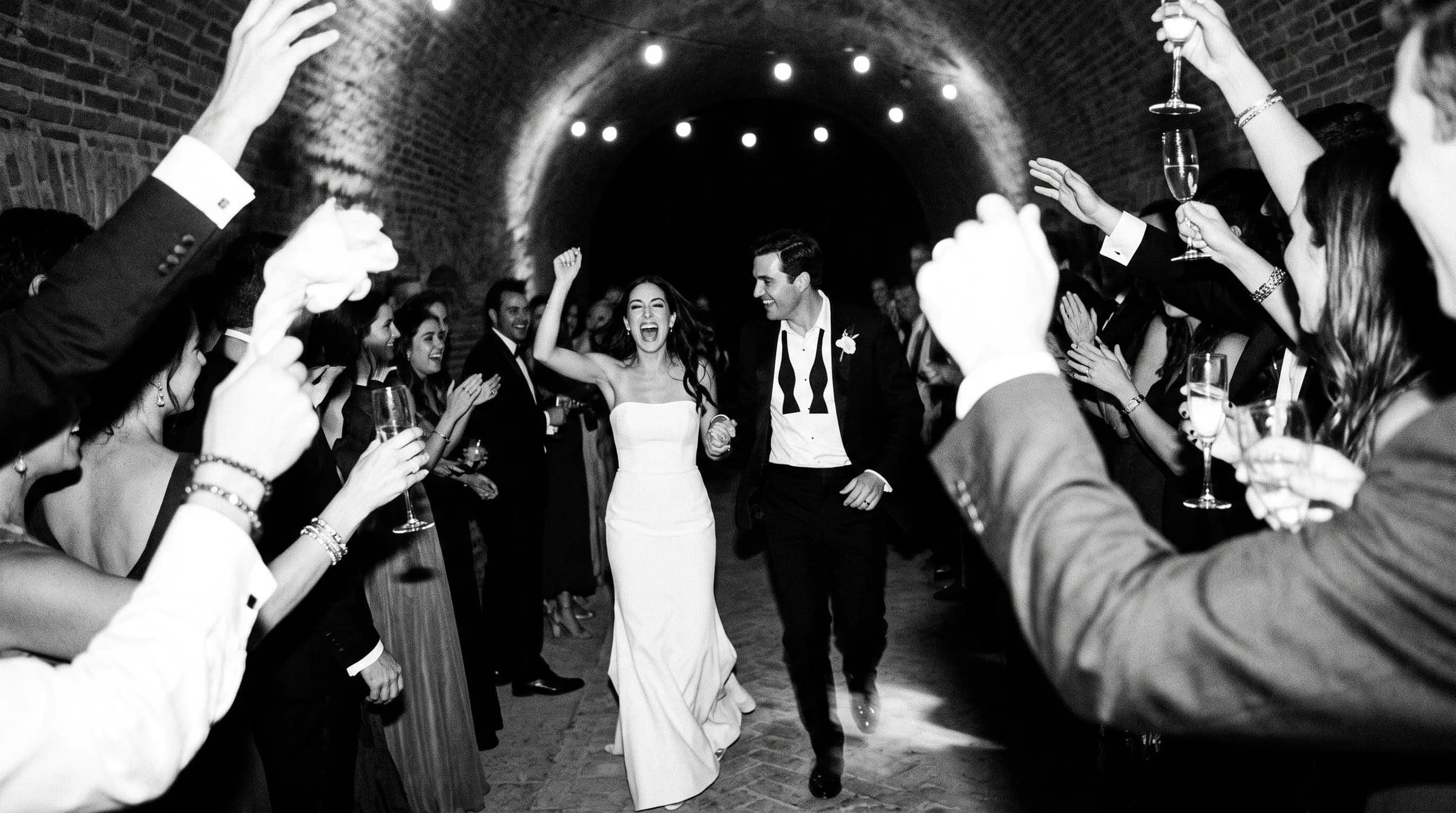 Novios saliendo entre invitados que aplauden en la bóveda de ladrillo de una hacienda, fotografía en blanco y negro